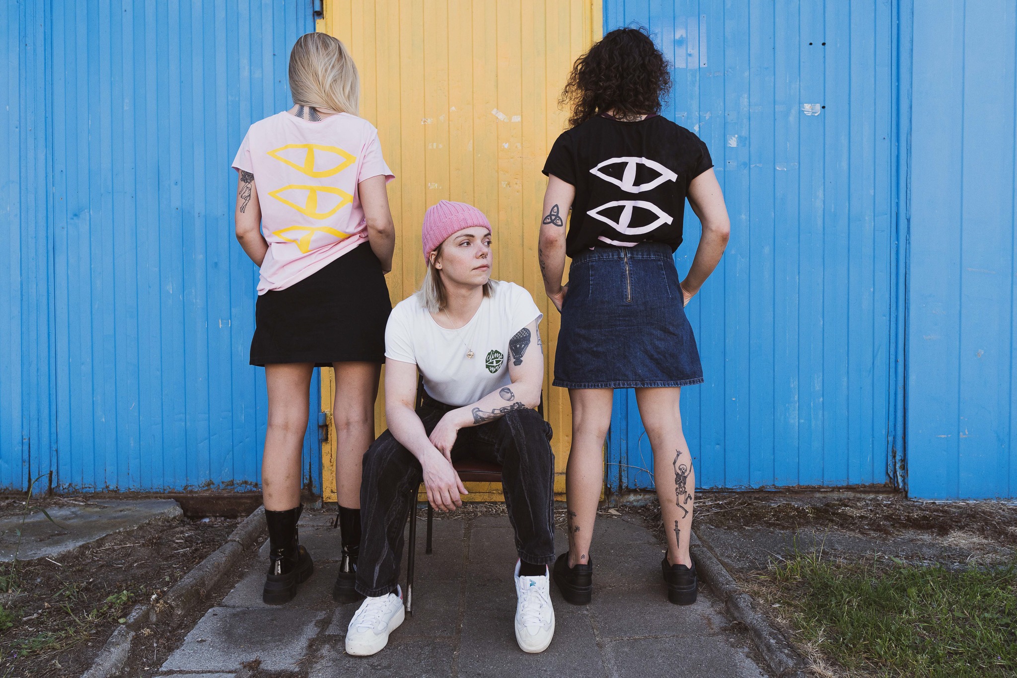 Trzy osoby prezentują ubrania z nadrukiem na tle niebiesko-żółtej ściany; dwie osoby stoją tyłem w koszulkach, jedna siedzi przodem na krześle w białej koszulce i różowej czapce.