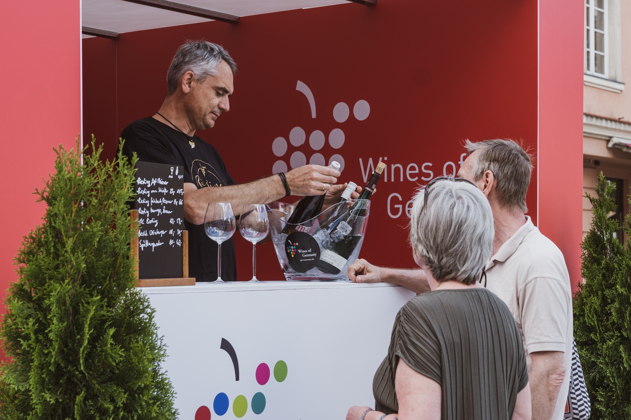 Sprzedawca nalewa wino z butelki trzymanej nad kubełkiem z lodem do kieliszka dla pary klientów przy stoisku z menu na tablicy i logiem Wines of Germany