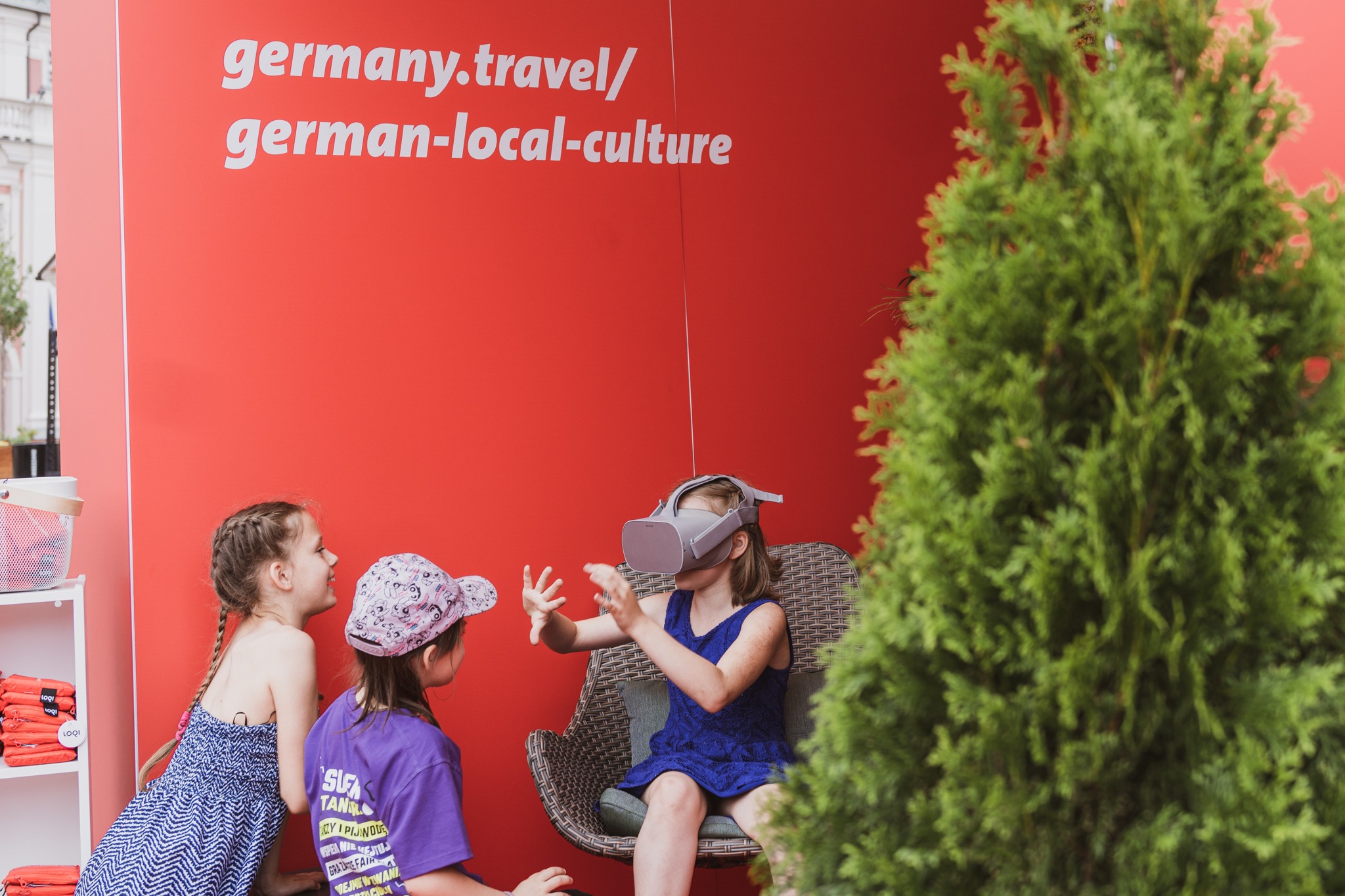Dwie dziewczynki obserwują koleżankę w niebieskiej sukience, która siedzi w wiklinowym fotelu i używa zestawu VR na tle czerwonej ściany z napisem germany.travel/german-local-culture oraz zielonego...