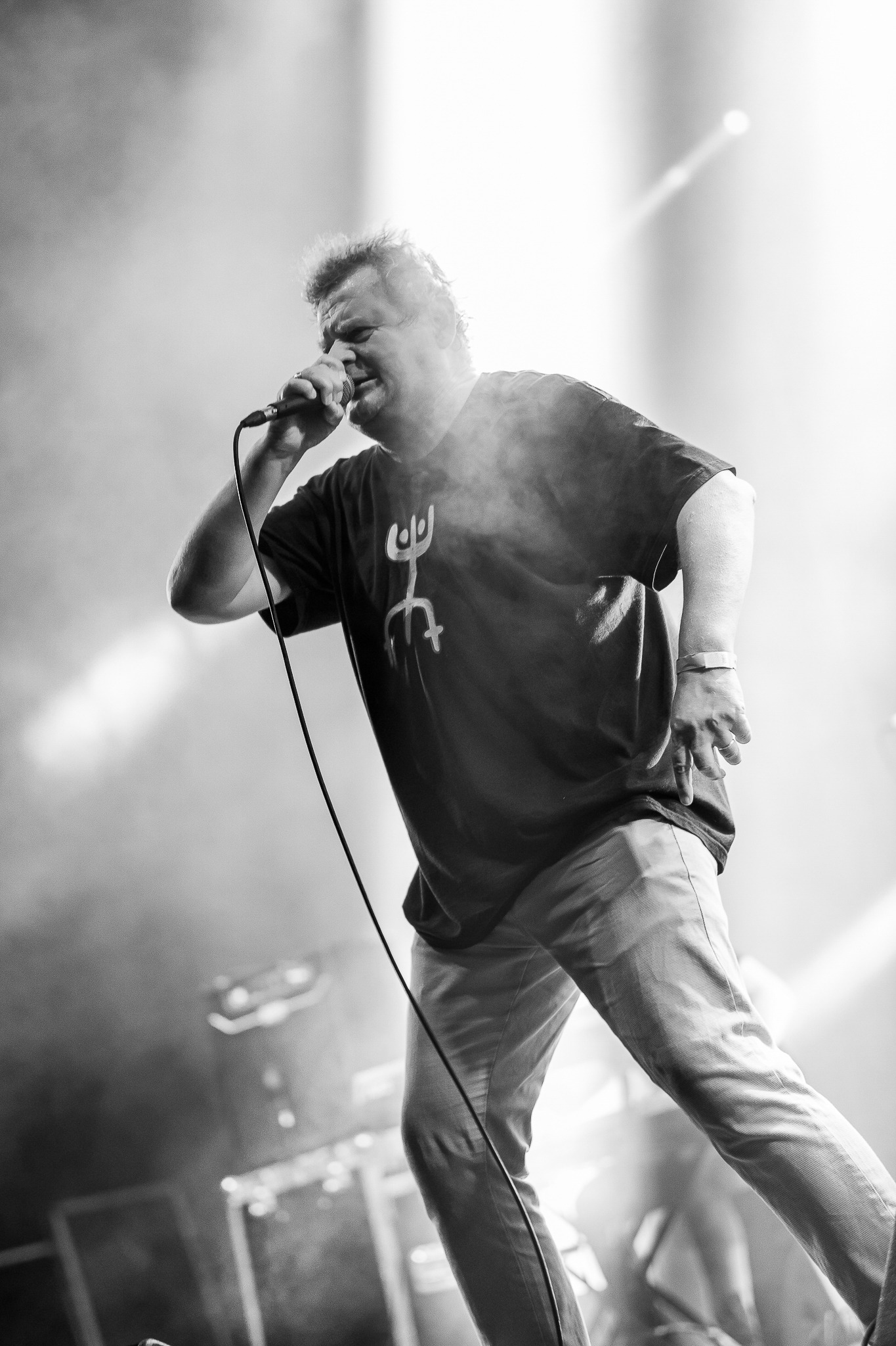 Czarno-białe zdjęcie wokalisty z mikrofonem podczas występu na scenie, oświetlonego reflektorami i otoczonego dymem.