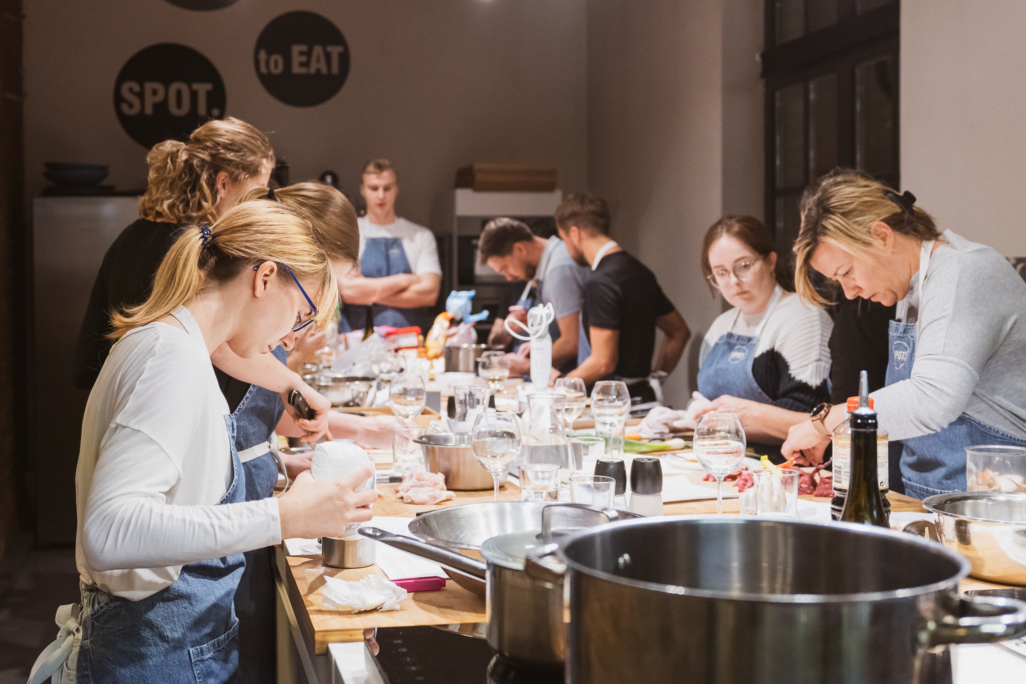 Grupa osób w fartuchach uczestniczy w warsztatach kulinarnych, przygotowując składniki na drewnianym stole, z widocznymi naczyniami, nożami i kieliszkami.