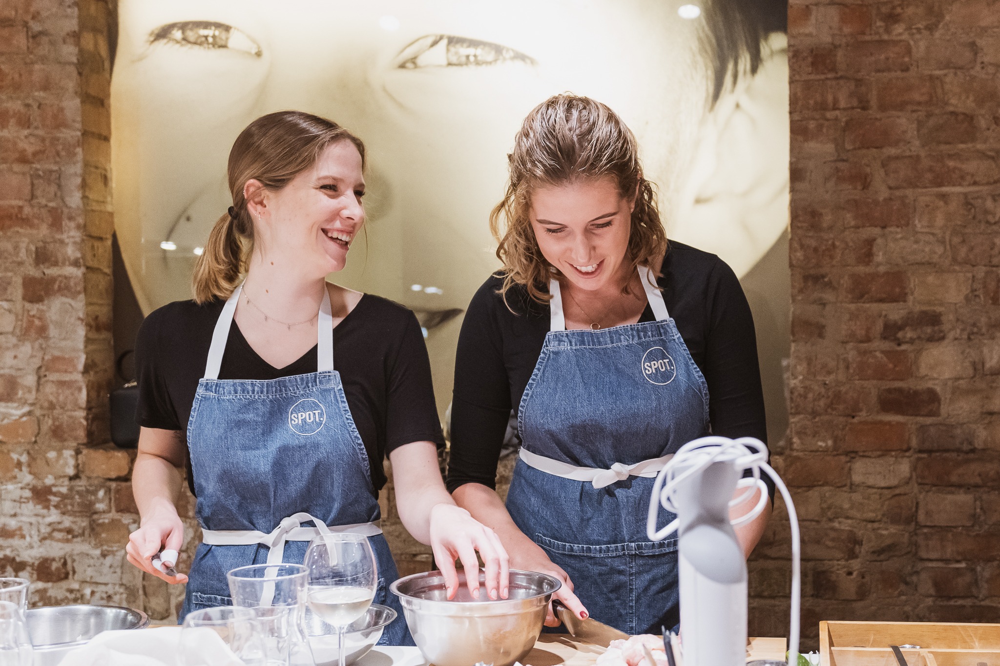 Dwie uśmiechnięte kobiety w dżinsowych fartuchach przygotowują posiłek w kuchni z ceglaną ścianą i artystyczną grafiką w tle, mieszając składniki w metalowej misce.