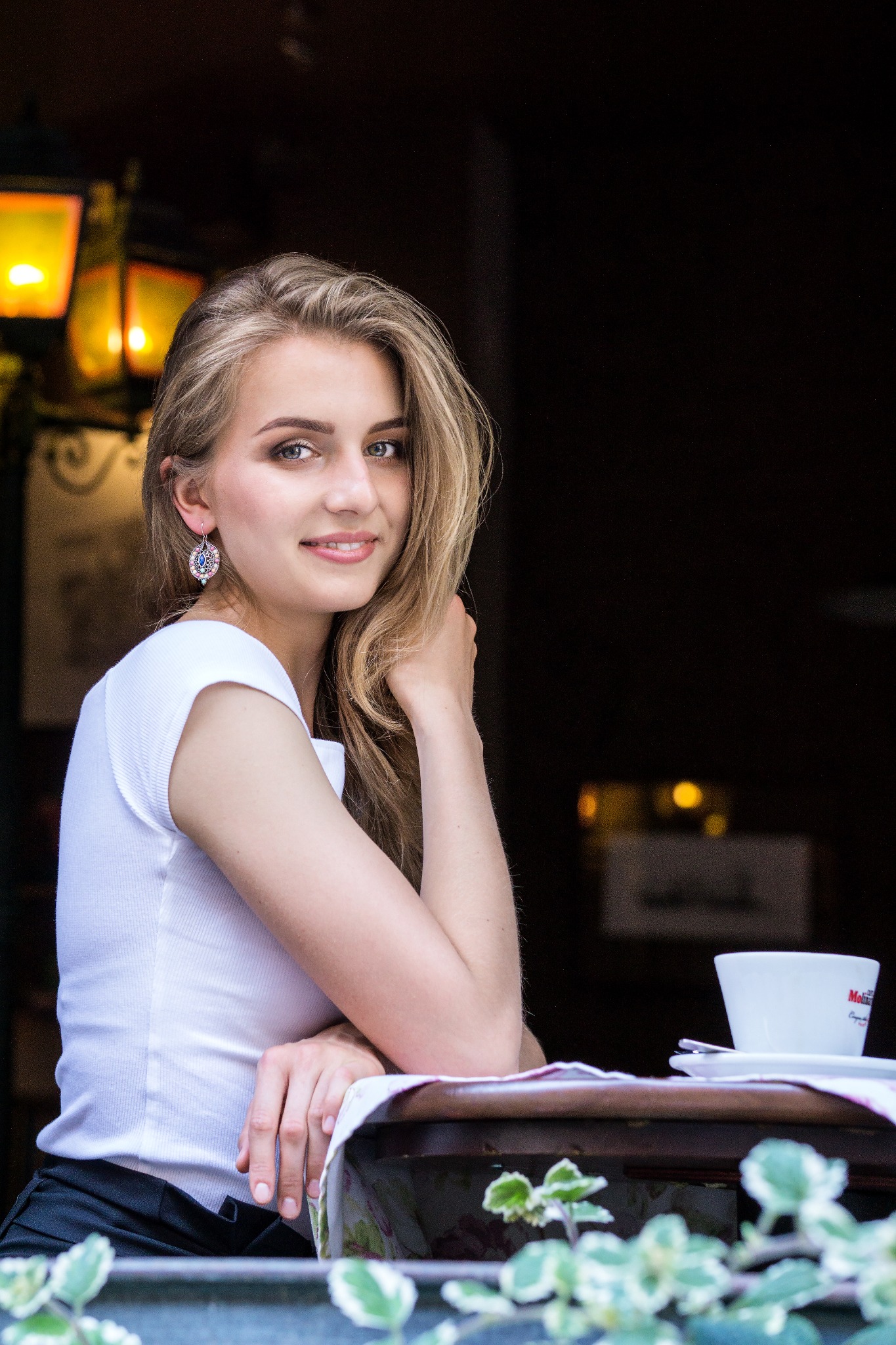 Portret młodej kobiety z blond włosami, opierającej się o stół z filiżanką kawy na zewnątrz kawiarni, oświetlonej ciepłym światłem latarni.