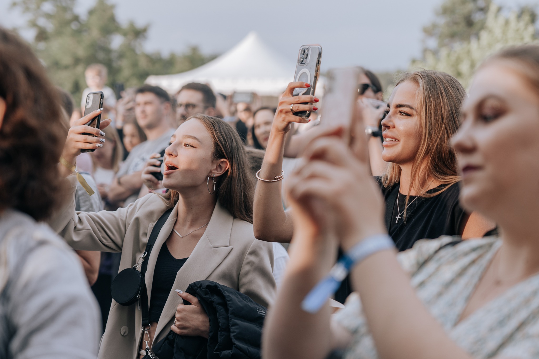 Grupa młodych ludzi, w tym dwie kobiety w centrum kadru, filmuje lub fotografuje coś smartfonami na tle białego namiotu i drzew, prawdopodobnie podczas wydarzenia plenerowego.