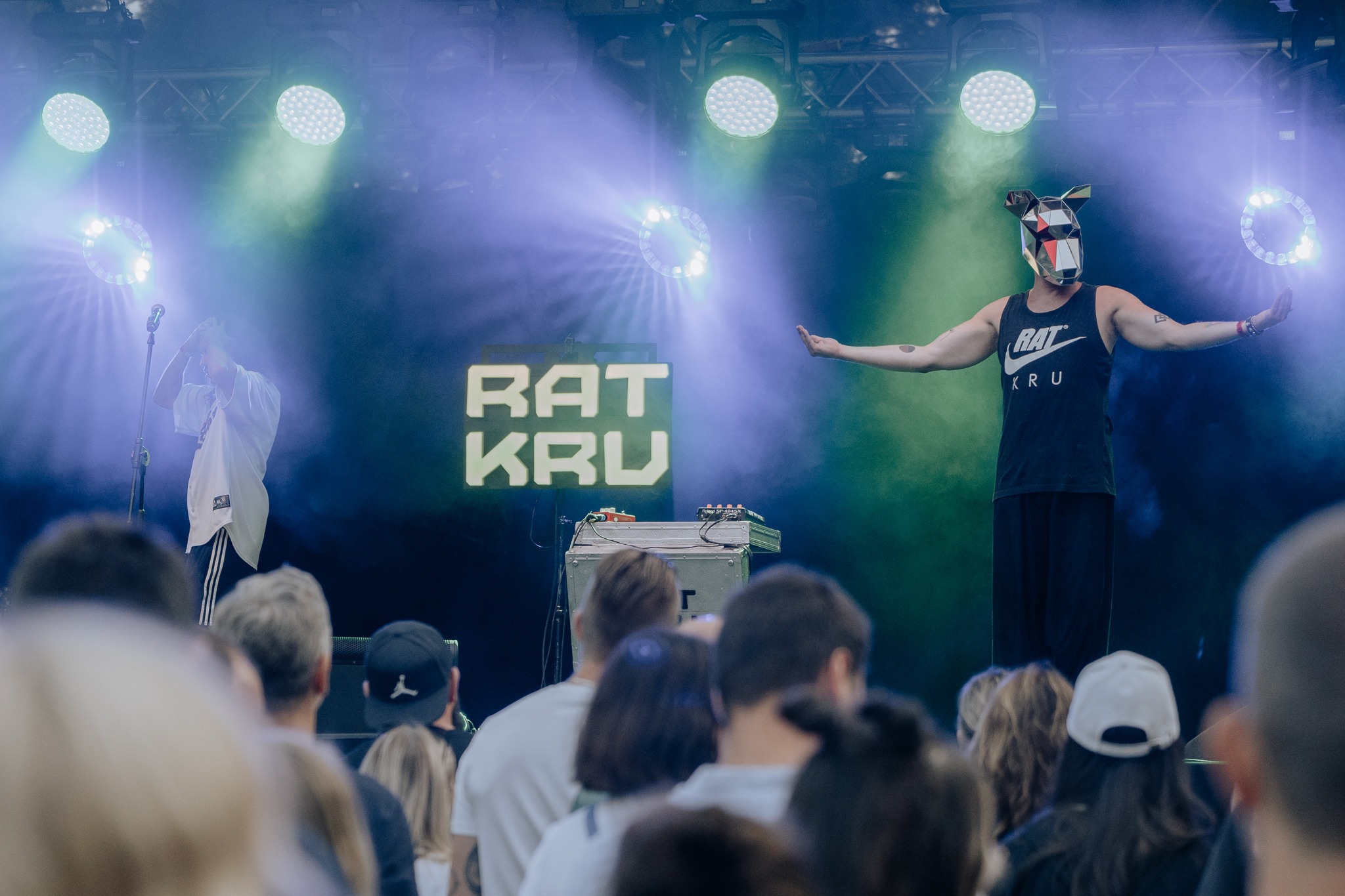 Koncert zespołu Rat Kru: mężczyzna w masce geometrycznej i koszulce bez rękawów stoi na scenie z rozłożonymi ramionami, za nim neonowy napis z nazwą zespołu i drugi muzyk przy mikrofonie, widownia...