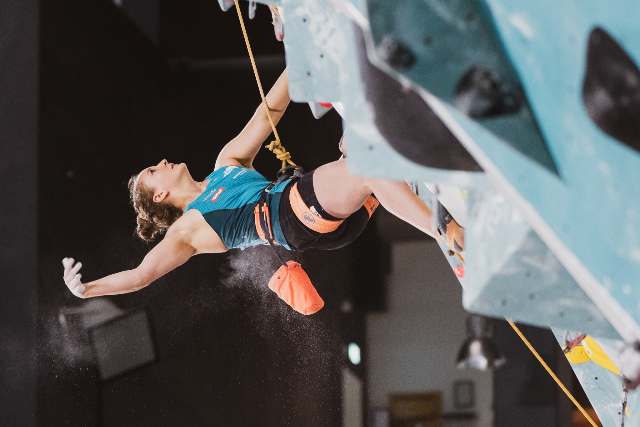 Kobieta wspinająca się na sztucznej ściance, w uprzęży i z magnezją, w dynamicznej pozycji z wyciągniętą ręką, widok z dołu.