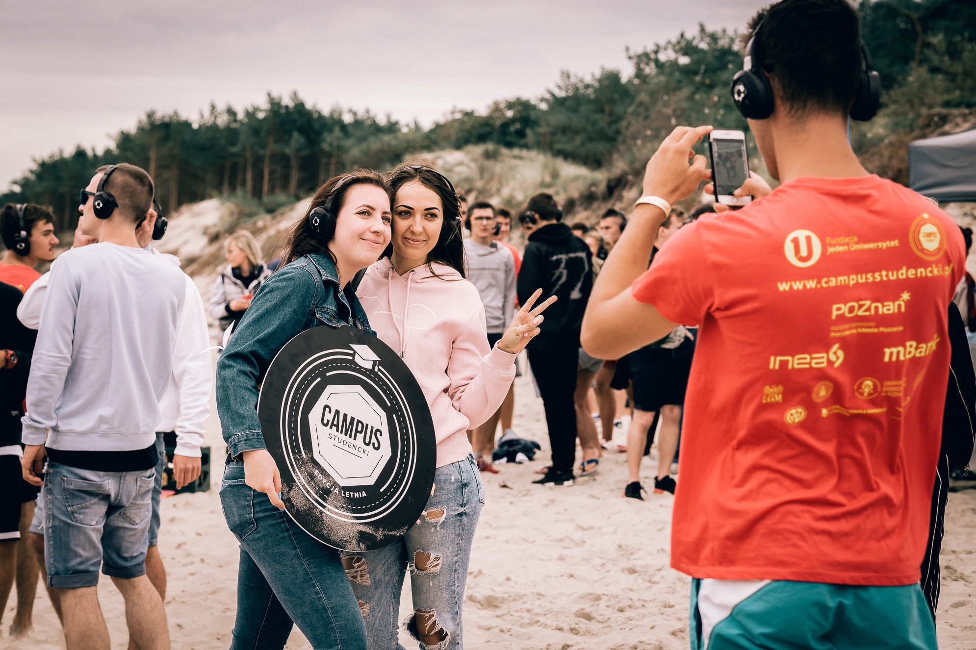 Dwie uśmiechnięte kobiety w słuchawkach pozują na plaży, trzymając okrągły znak 'Campus Studencki Edycja Letnia', podczas gdy mężczyzna w czerwonej koszulce robi im zdjęcie telefonem, a w tle widać...