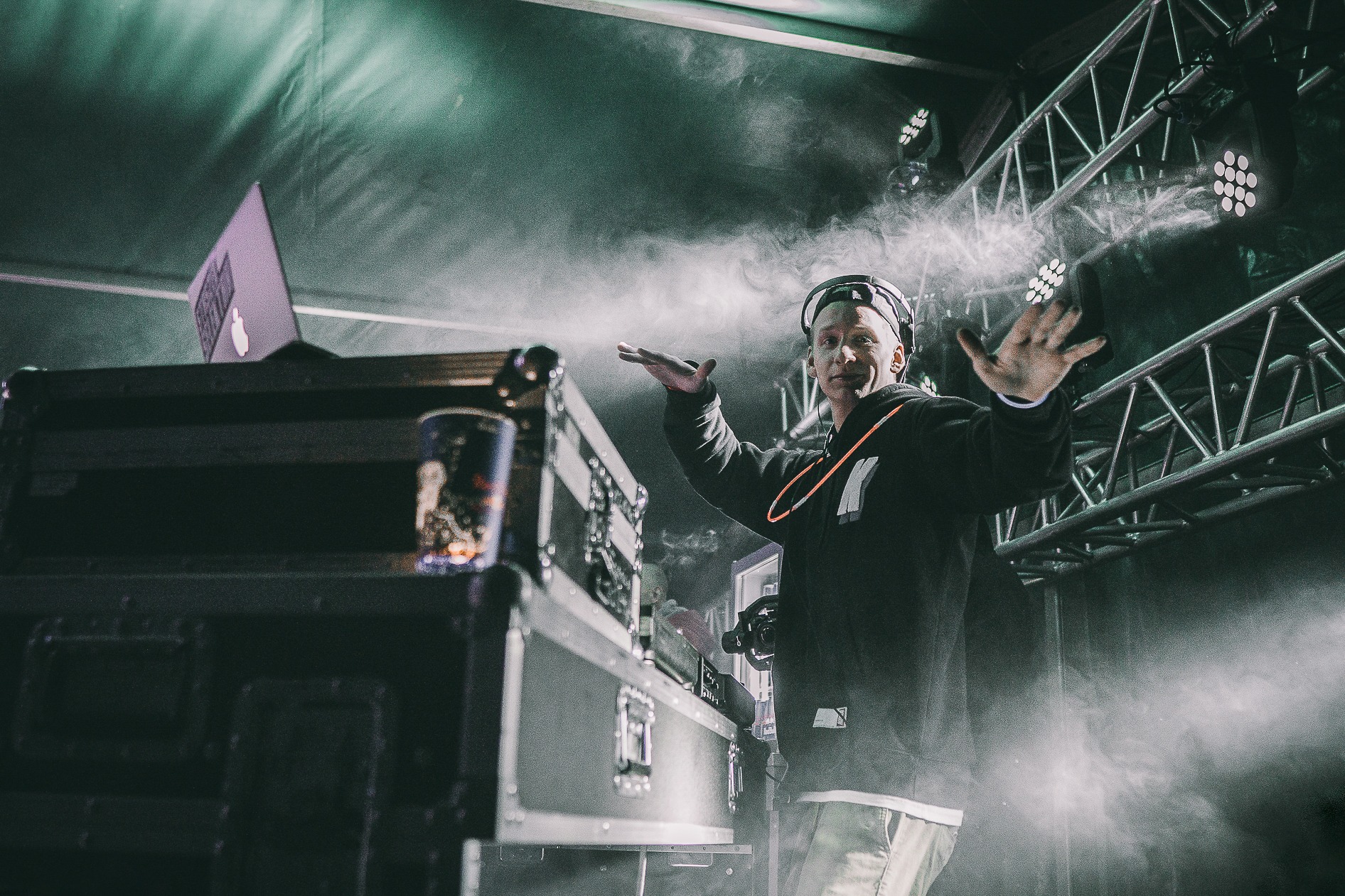 DJ w słuchawkach, z rękami uniesionymi w geście interakcji z publicznością, oświetlony scenicznie z efektami dymu, laptop i napój na stole DJ-skim, metalowa konstrukcja sceniczna w tle.