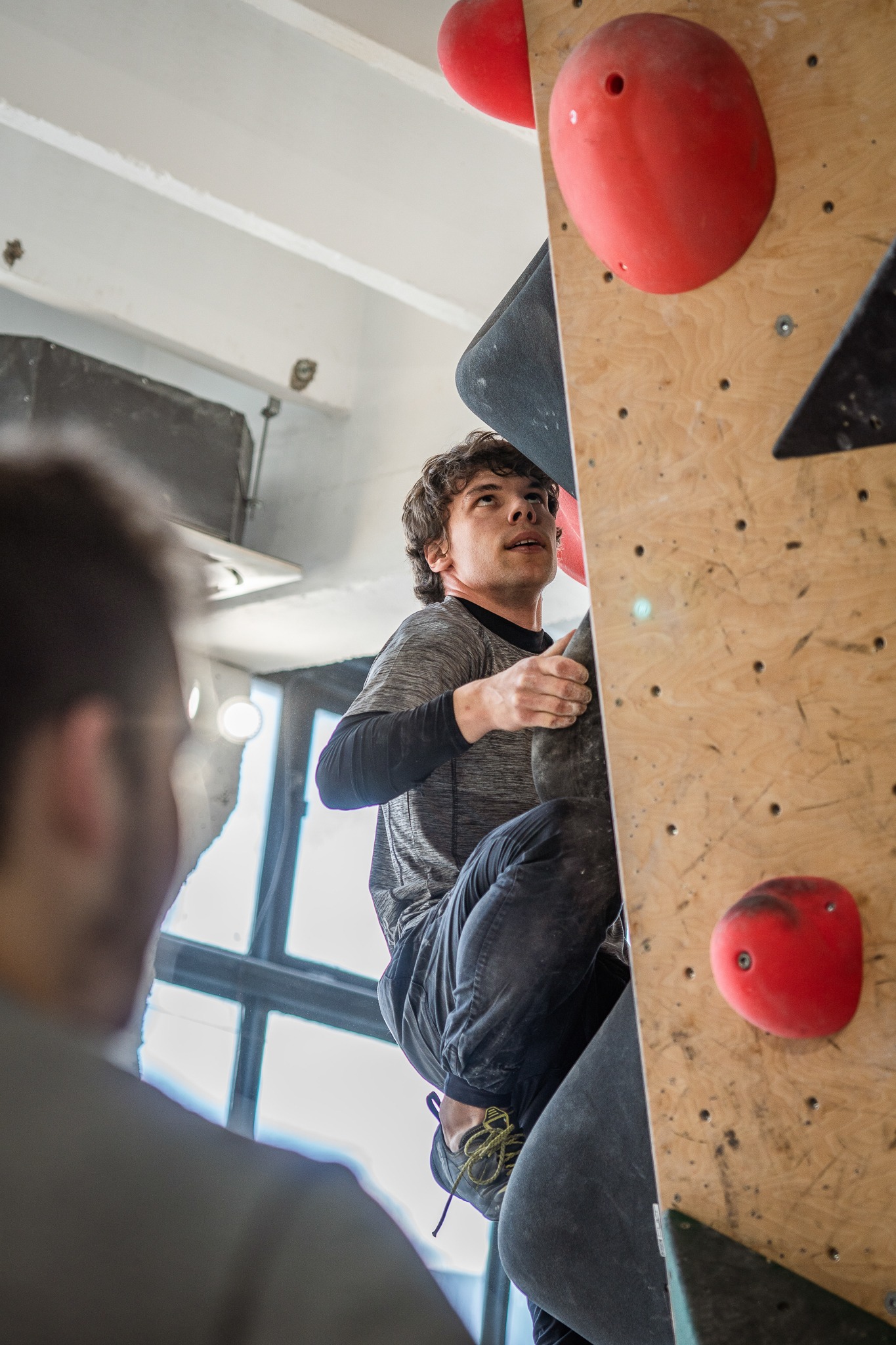 Młody mężczyzna w szarej koszulce i czarnych spodniach wspina się na ścianę wspinaczkową z czerwonymi i czarnymi chwytami, w tle widoczna sylwetka innej osoby.