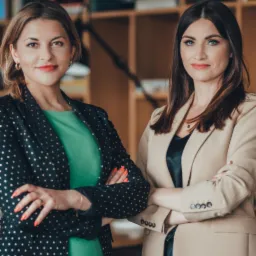 Portret dwóch pewnych siebie kobiet w eleganckich marynarkach na tle regału z książkami, skrzyżowane ręce, profesjonalny wizerunek.