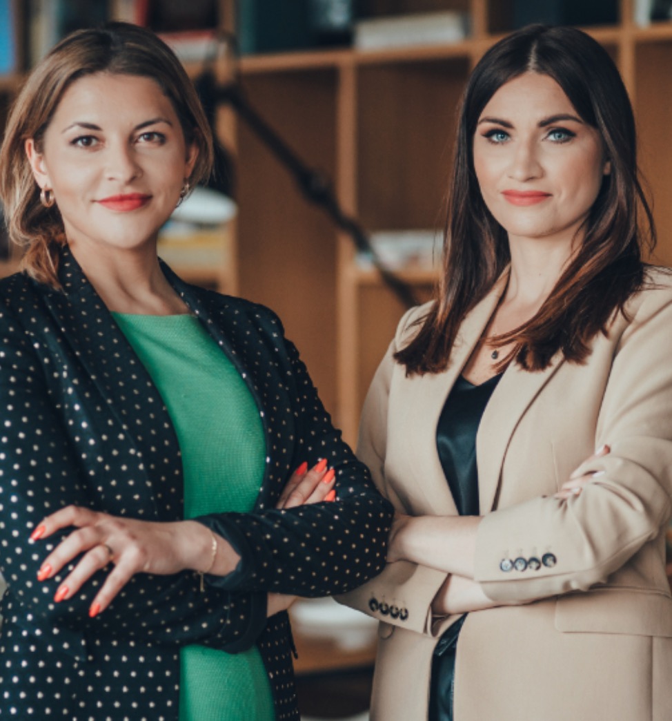 Portret dwóch pewnych siebie kobiet w eleganckich marynarkach na tle regału z książkami, skrzyżowane ręce, profesjonalny wizerunek.