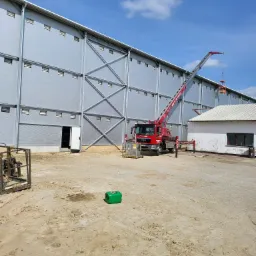 Szara hala magazynowa z konstrukcją stalową, czerwony podnośnik koszowy z wysięgnikiem sięgającym dachu, piaszczysty teren budowy.