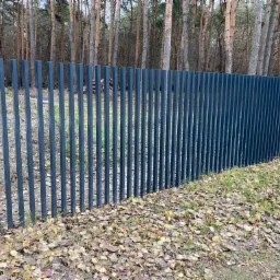 Nowoczesne ogrodzenie panelowe z pionowych, grafitowych profili na tle lasu iglastego i opadłych liści.