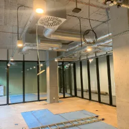 Wnętrze biurowe w trakcie prac wykończeniowych: widoczne instalacje pod sufitem, oświetlenie, wentylacja, drabina na podłodze zabezpieczonej folią, szklane ścianki działowe.