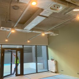 Vladyslav Ruban - Wnętrze w stanie deweloperskim z widocznymi instalacjami pod sufitem, pracownikiem w kamizelce otwierającym szklane drzwi oraz niezabezpieczonymi puszkami elektrycznymi na ścianie.