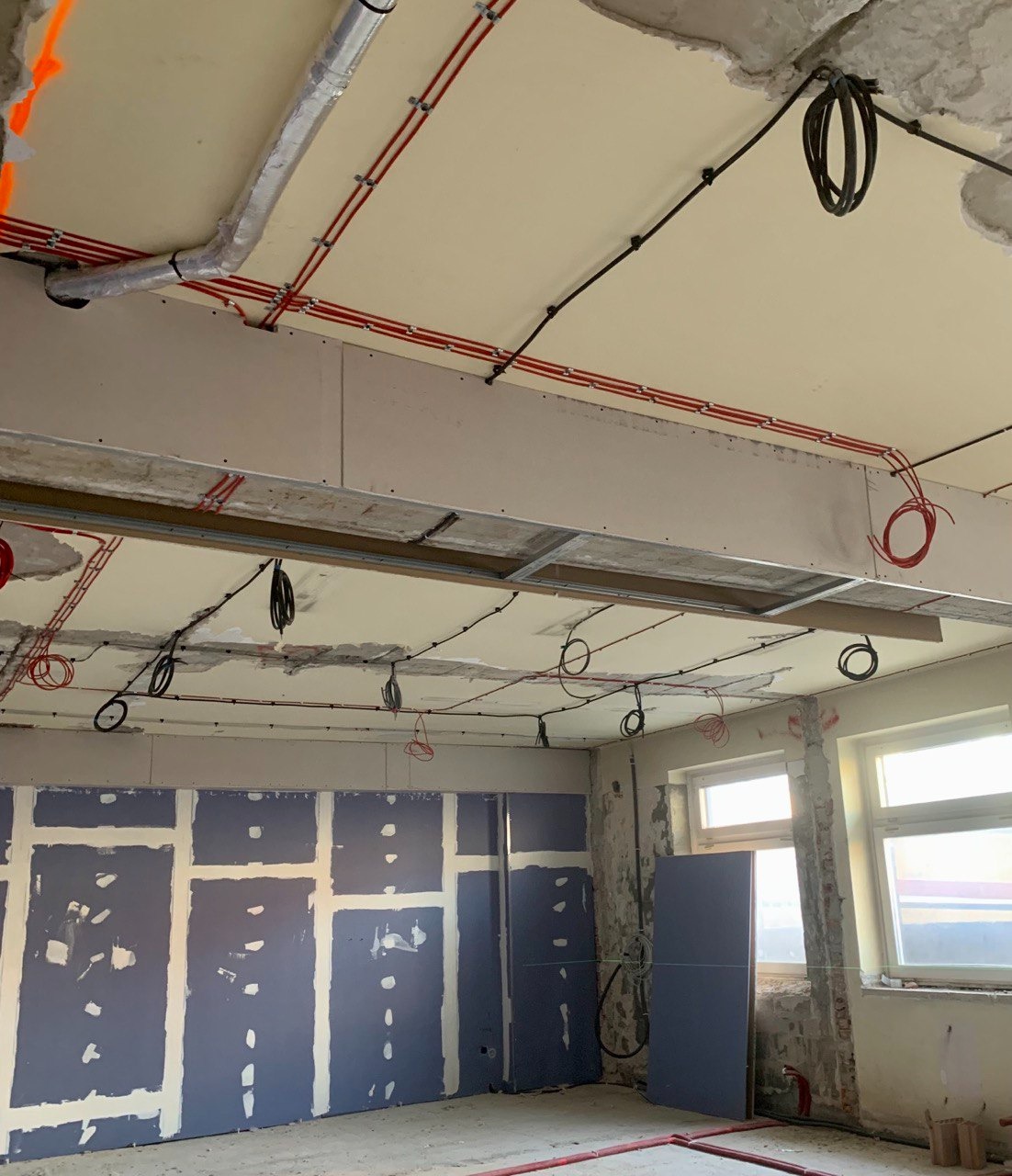 Wnętrze w trakcie remontu z widocznymi instalacjami: czerwone i czarne kable poprowadzone po suficie i ścianach, częściowo zakryte płytami gipsowo-kartonowymi, widoczne elementy wentylacji i okna.