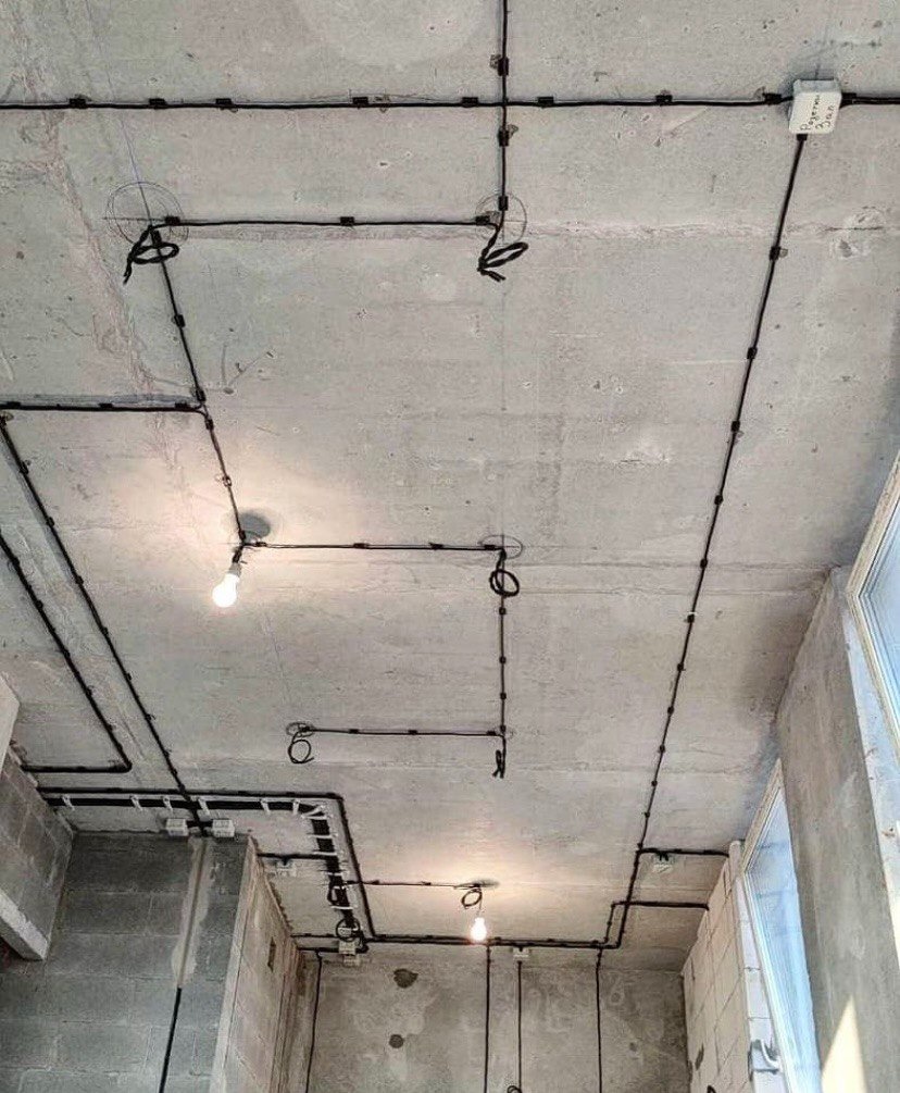 Surowy betonowy sufit z widocznymi, czarnymi przewodami elektrycznymi przymocowanymi klamrami, żarówka wkręcona tymczasowo w oprawkę, widoczne okno z prawej strony.