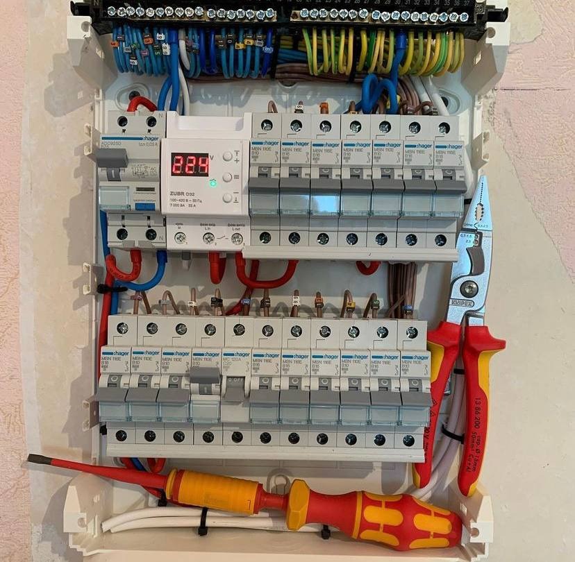 Otwarta skrzynka elektryczna z zamontowanymi bezpiecznikami Hager, przewodami w różnych kolorach i narzędziami: kombinerki Knipex oraz śrubokręt z żółto-czerwoną rękojeścią.