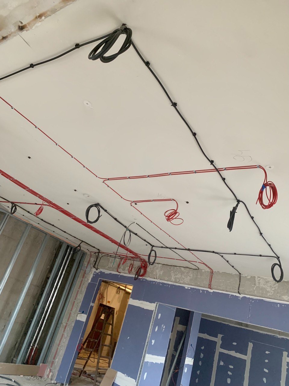 Rozprowadzone przewody elektryczne w kolorze czerwonym i czarnym, zamocowane do sufitu w surowym stanie, widoczne metalowe profile ścian działowych i drabina w tle.