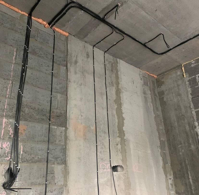 Surowe wnętrze z betonu z widocznymi, czarnymi przewodami elektrycznymi, przymocowanymi do ścian i sufitu za pomocą białych uchwytów. Widoczne ślady ołówka na ścianach.