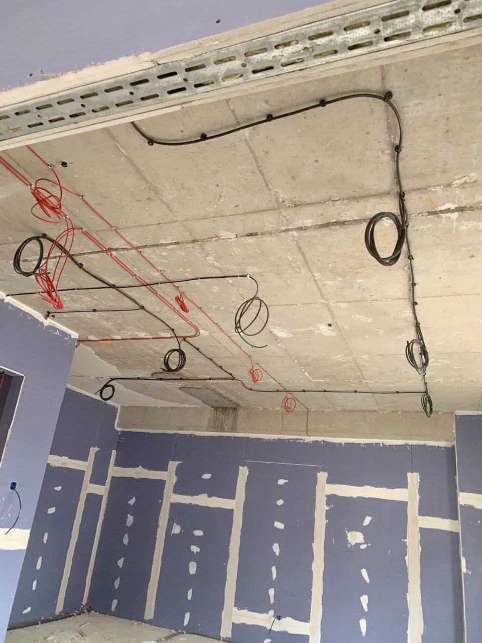 Przewody elektryczne w kolorze czerwonym i czarnym prowadzone po suficie podwieszanym w trakcie montażu, widoczne metalowe korytko kablowe i ściany z płyt gipsowo-kartonowych.