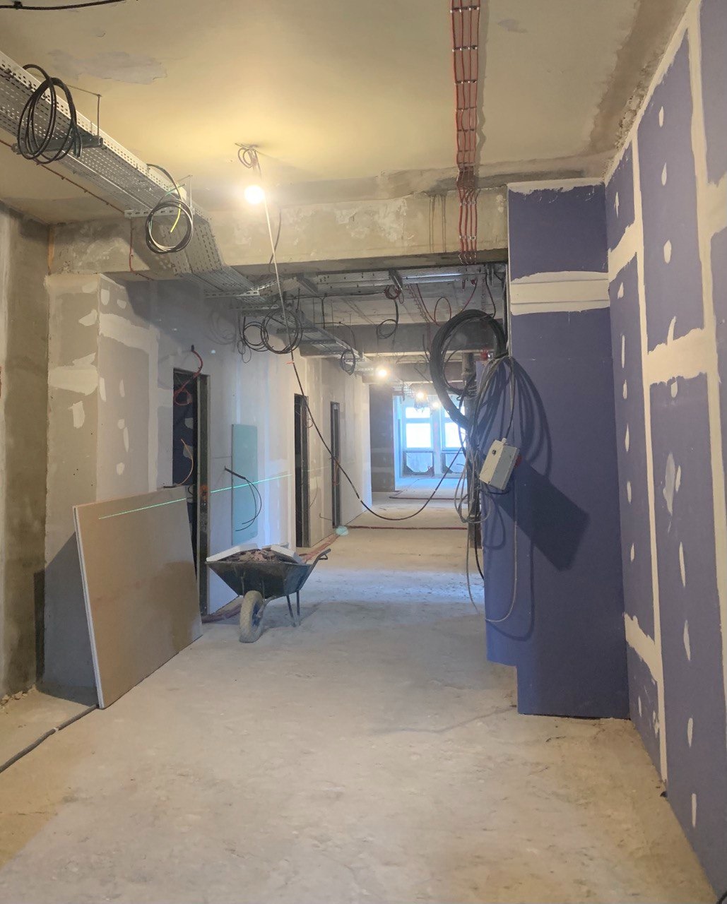 Korytarz w trakcie remontu z widocznymi przewodami elektrycznymi na suficie, płytami gipsowymi na ścianach i taczką z gruzem.