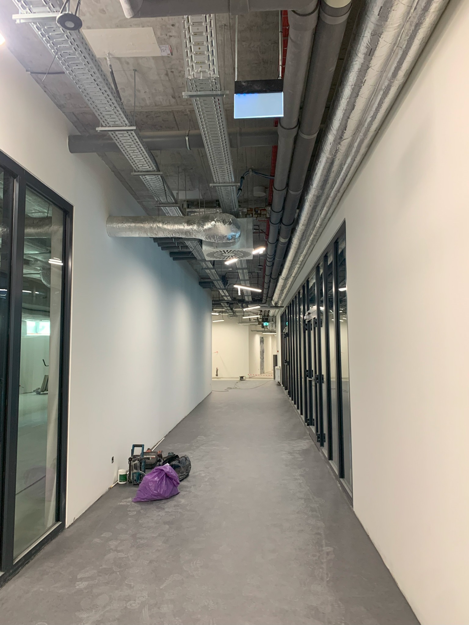 Korytarz w biurowcu w trakcie prac instalacyjnych: widoczne koryta kablowe pod sufitem, rury wentylacyjne, narzędzia i worek na podłodze.