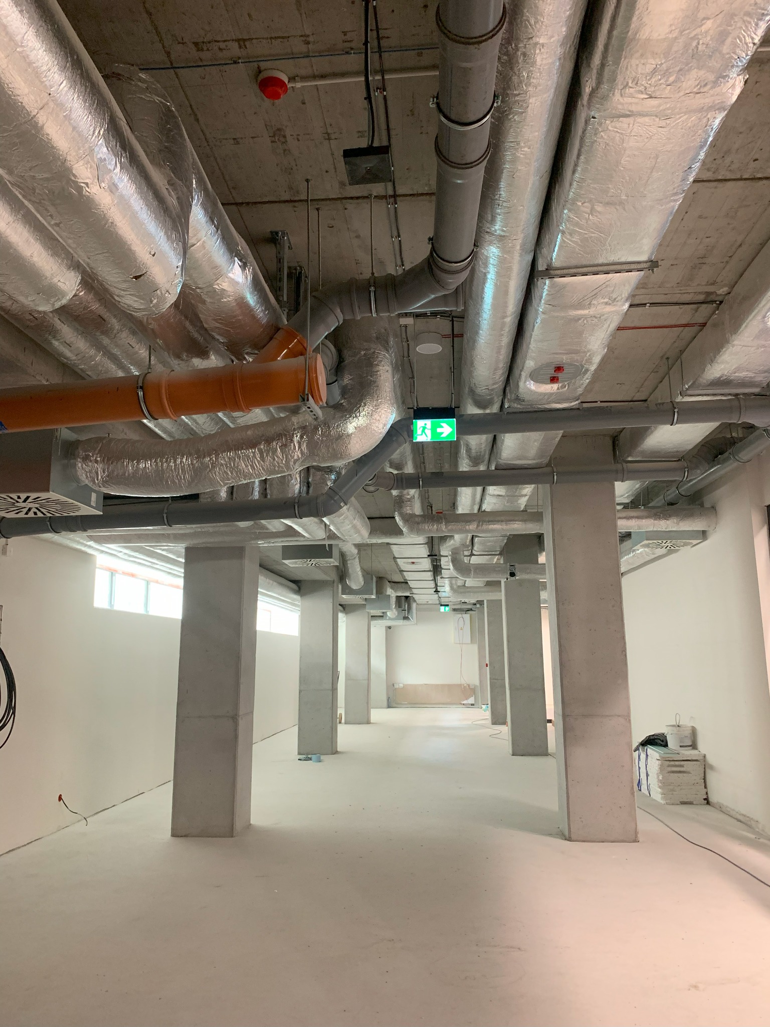 Widok sufitu w surowym stanie, z licznymi rurami i przewodami wentylacyjnymi, niektóre owinięte srebrną folią izolacyjną, oraz zielonym znakiem ewakuacyjnym, w tle widoczne kolumny podtrzymujące...