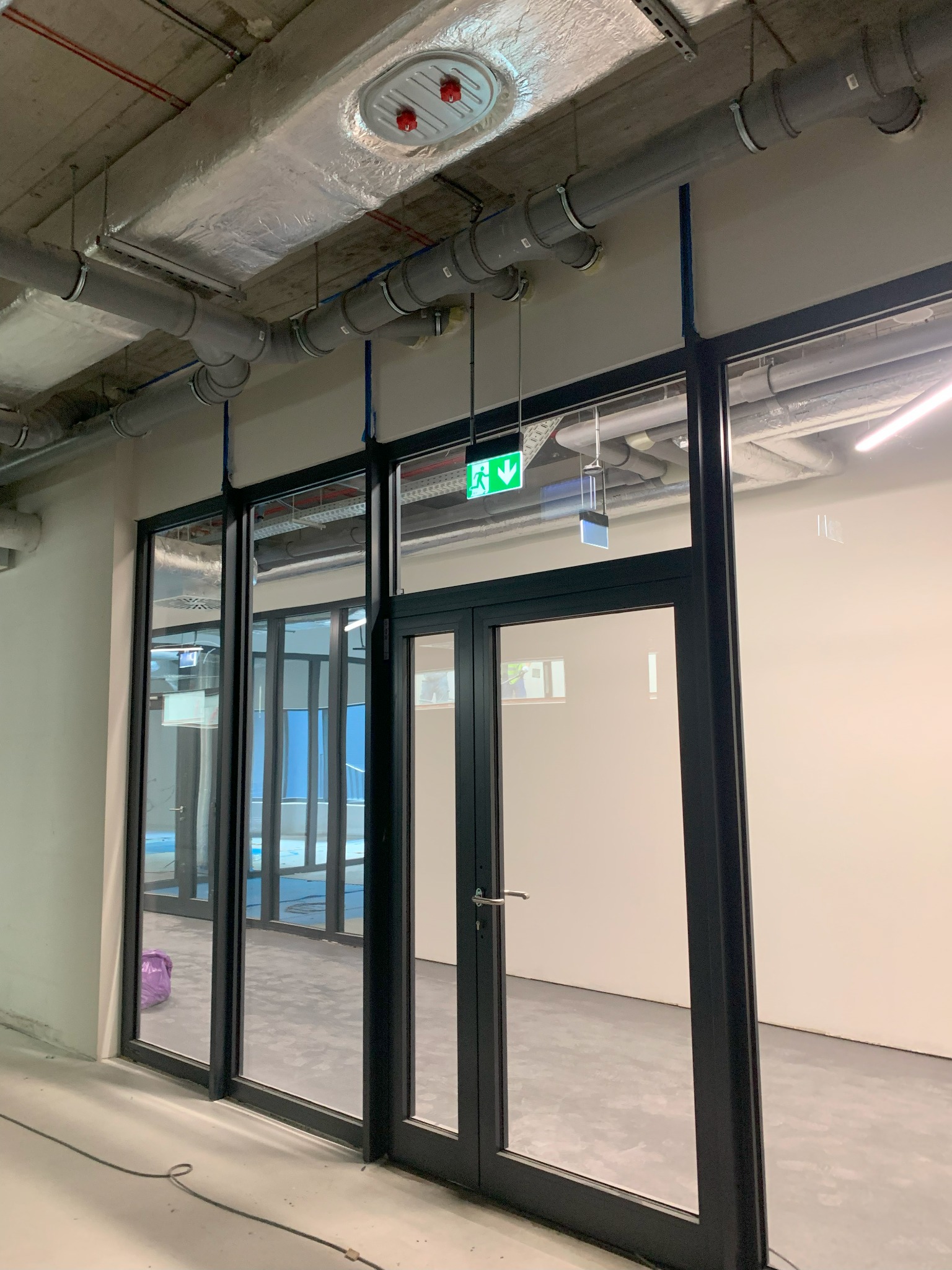 Szklane drzwi ewakuacyjne z czarną ramą w surowym wnętrzu biurowym, widoczne rury wentylacyjne i system przeciwpożarowy pod sufitem.