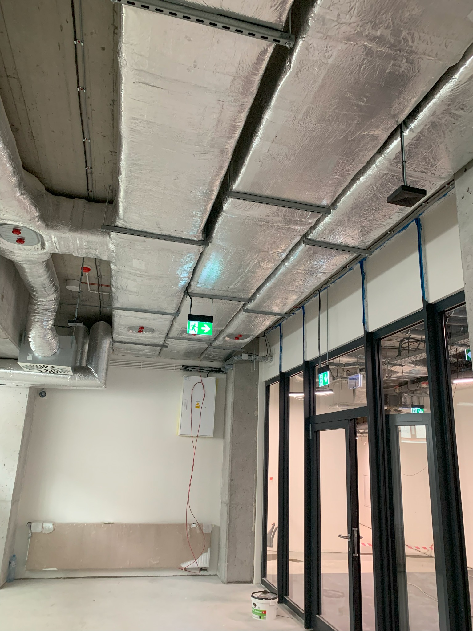 Wnętrze w trakcie wykańczania: widoczne rury wentylacyjne zaizolowane folią aluminiową pod sufitem, okablowanie, lampa awaryjna z zieloną strzałką wskazującą wyjście, oraz szklane drzwi.