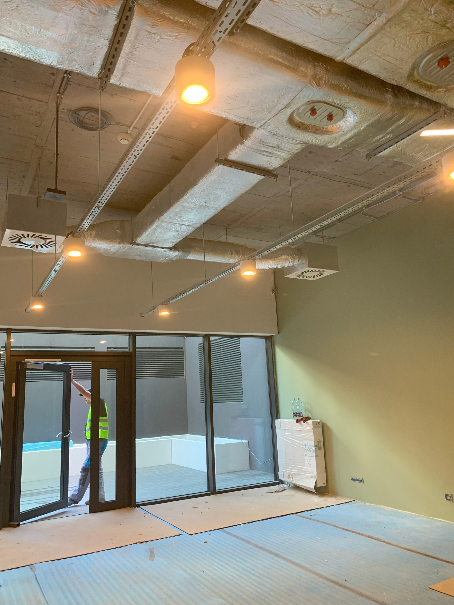 Wnętrze w stanie deweloperskim z widocznymi instalacjami pod sufitem, pracownikiem w kamizelce otwierającym szklane drzwi oraz niezabezpieczonymi puszkami elektrycznymi na ścianie.