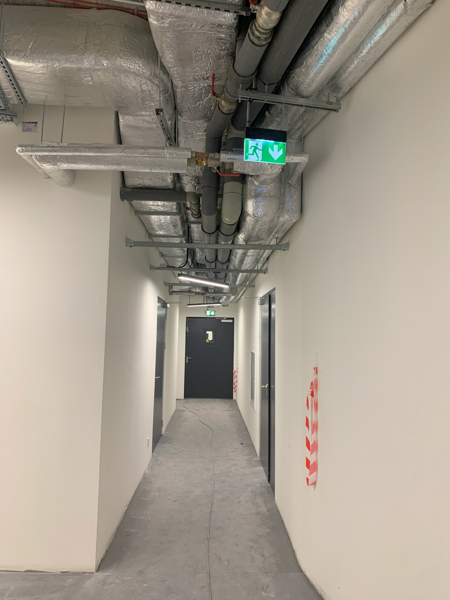 Korytarz w budynku z widocznymi instalacjami pod sufitem, w tym rurami w izolacji, przewodami i zielonym znakiem ewakuacyjnym ze strzałką w dół.