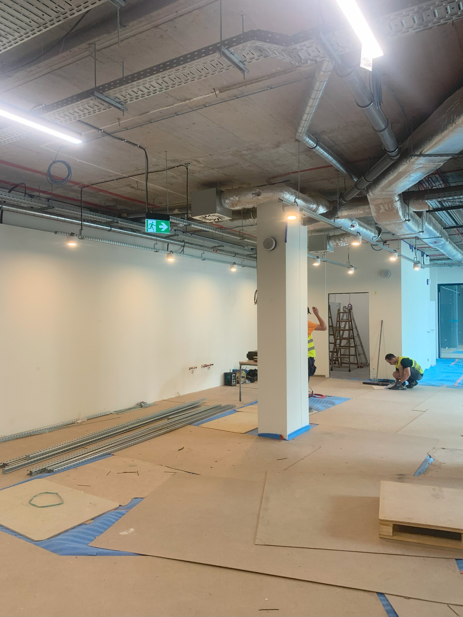 Prace instalacyjne w nowym lokalu w Krakowie: widoczne koryta kablowe pod sufitem, zamontowane oświetlenie punktowe, pracownicy w kamizelkach odblaskowych oraz przygotowane materiały i narzędzia...