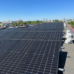 Eco Energia Anna Lisiecka - Instalacja fotowoltaiczna na płaskim dachu budynku z widocznymi inwerterami i elementami montażowymi, na tle miejskiego krajobrazu pod błękitnym niebem.