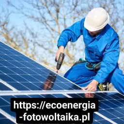 Eco Energia Anna Lisiecka - Pracownik w niebieskim kombinezonie i białym kasku montuje panele fotowoltaiczne za pomocą wkrętarki na dachu w słoneczny dzień. Widoczne drzewa w tle.