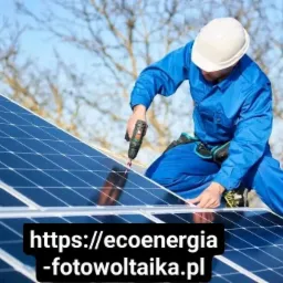 Pracownik w niebieskim kombinezonie i białym kasku montuje panele fotowoltaiczne za pomocą wkrętarki na dachu w słoneczny dzień. Widoczne drzewa w tle.