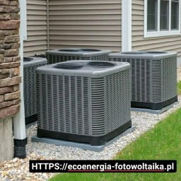 Eco Energia Anna Lisiecka - Cztery szare jednostki zewnętrzne klimatyzacji ustawione obok domu z brązową elewacją, widoczne na tle trawnika i kamiennego podłoża.