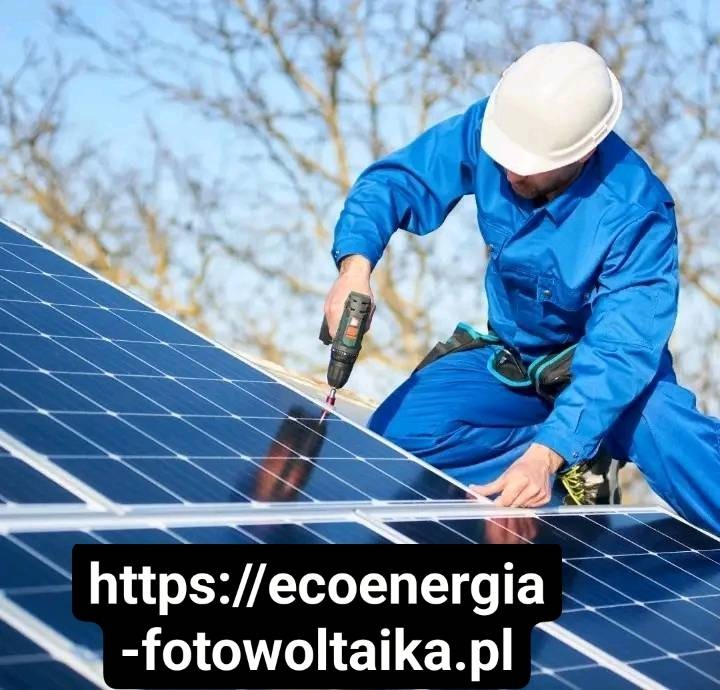 Pracownik w niebieskim kombinezonie i białym kasku montuje panele fotowoltaiczne za pomocą wkrętarki na dachu w słoneczny dzień. Widoczne drzewa w tle.