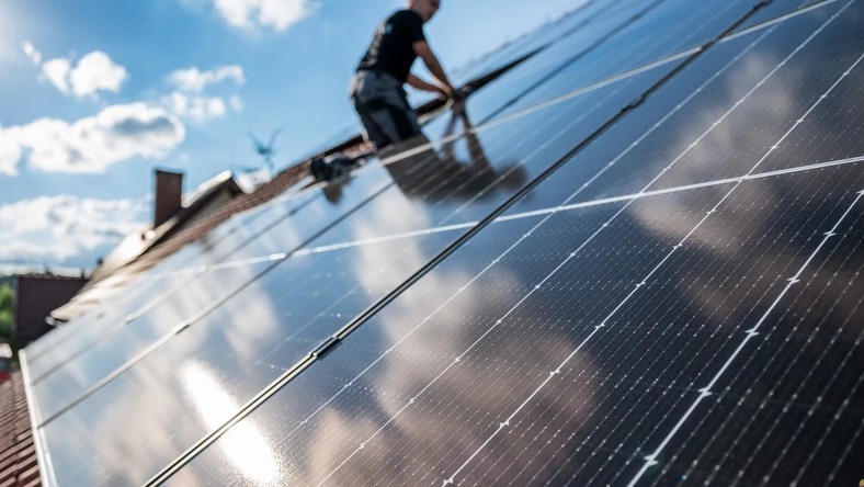 Instalacja paneli słonecznych na dachu domu z ceglaną dachówką, widok z dołu na instalatora w czarnej koszulce na tle błękitnego nieba z chmurami.