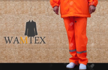 Osoba w jaskrawo pomarańczowym komplecie odzieży ochronnej, składającym się z kurtki i spodni z odblaskowymi paskami, stojąca na tle ściany z wzorem płytek. Na pierwszym planie widoczne logo firmy.