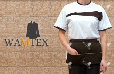 Kobieta w białej koszulce z brązowym paskiem i krótkim, brązowym fartuchu z kieszenią, pozuje na tle ściany z płytek. Na zdjęciu widoczne logo firmy odzieżowej.