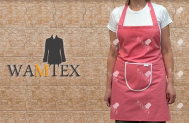Różowy fartuch roboczy z białą lamówką i kieszenią, prezentowany na modelce na tle ściany z płytek. Widoczne logo WAMTEX.