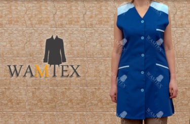 Niebieski fartuch roboczy z jasnoniebieskimi wstawkami na ramionach i kieszeniach, zapinany na guziki, prezentowany na modelce na tle ceglanej ściany. Widoczne logo WAMTEX.