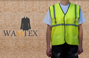 Osoba w żółtej kamizelce odblaskowej z czarnym obszyciem i szarymi pasami na tle ściany z wzorem płytek, widoczne logo firmy Wamtex w lewym dolnym rogu.