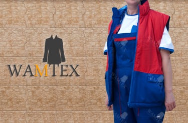 Osoba w niebieskim kombinezonie roboczym z czerwono-granatową ocieplaną kamizelką na tle ściany z imitacji kamienia, widoczne logo firmy WAMTEX.