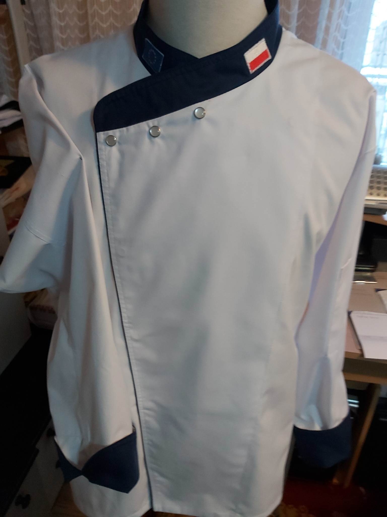 Biała bluza kucharska z granatowymi wykończeniami, widoczne naszywki z flagą Polski i Unii Europejskiej, zapinana na srebrne guziki, na manekinie.