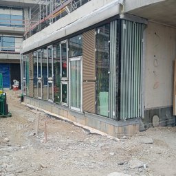 LK SP. ZO. O - Wnętrze budynku w stanie surowym zamkniętym z zamontowanymi oknami fasadowymi i lamelami pionowymi, widok na plac budowy z gruzem i rusztowaniami w tle.