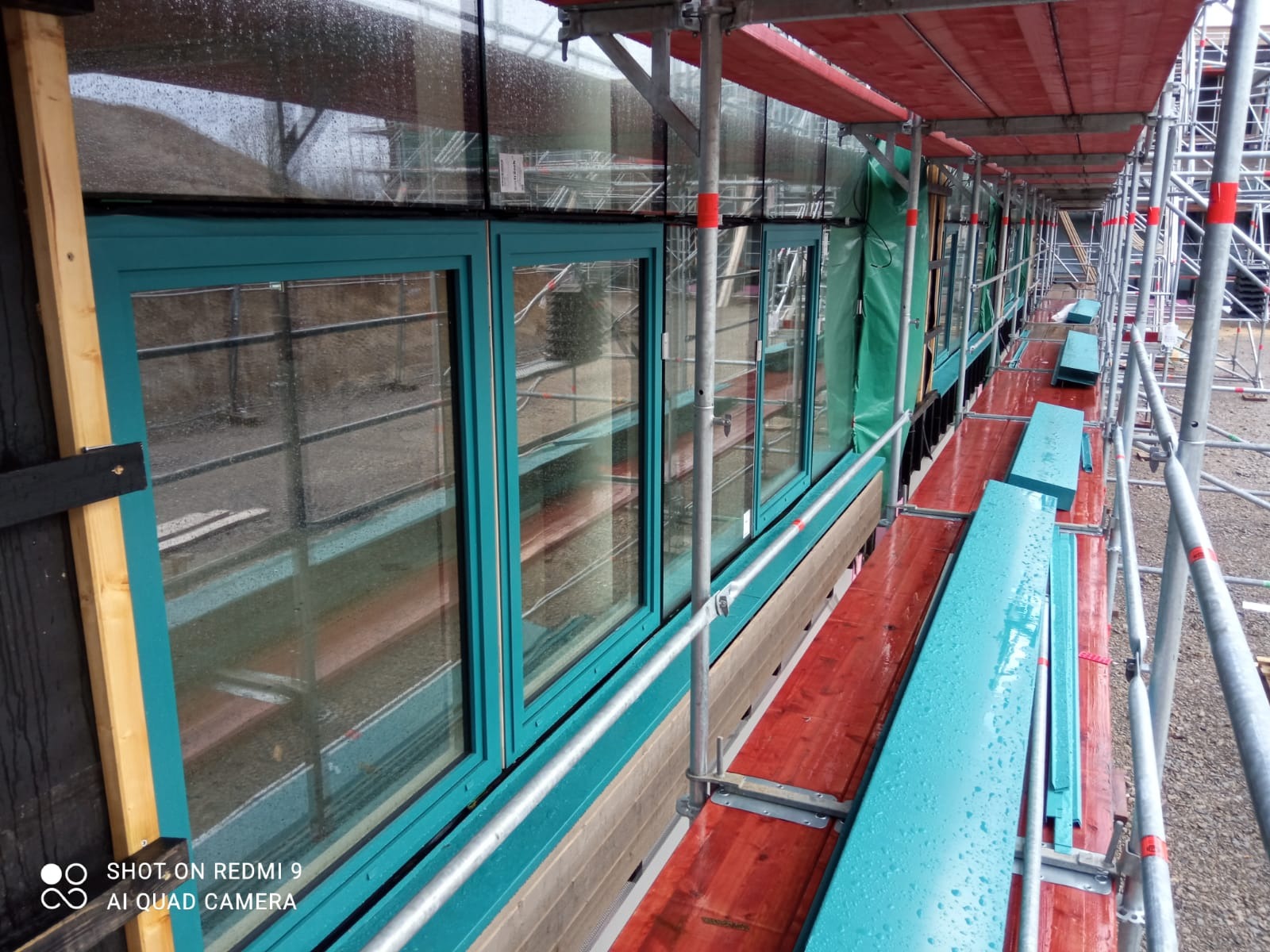 Montaż okien z turkusowymi ramami na elewacji budynku, widoczne rusztowanie, deszczowa pogoda.