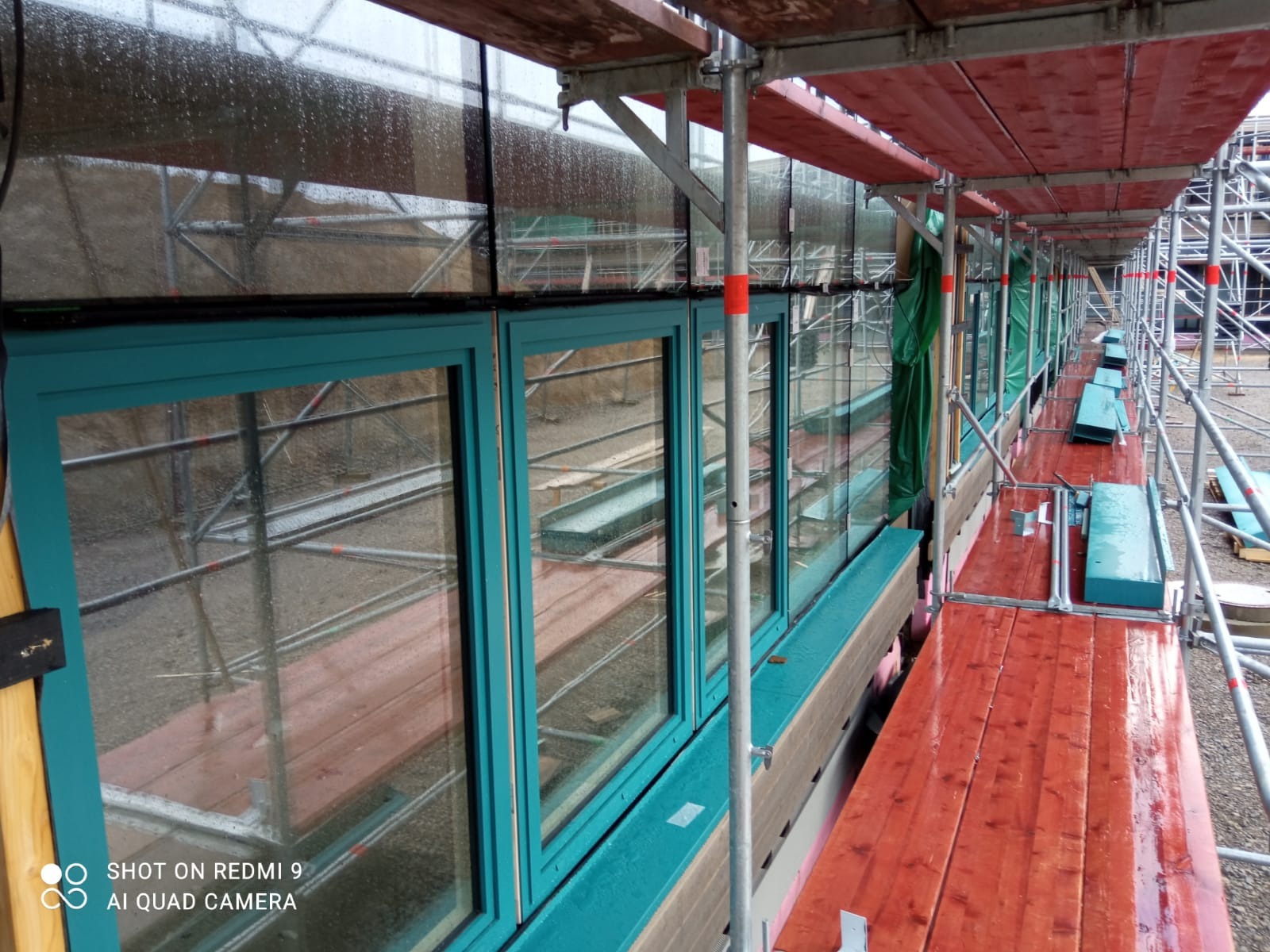 Montaż okien w kolorze turkusowym na budowie, widoczne rusztowanie i zabezpieczenia, deszczowa pogoda.