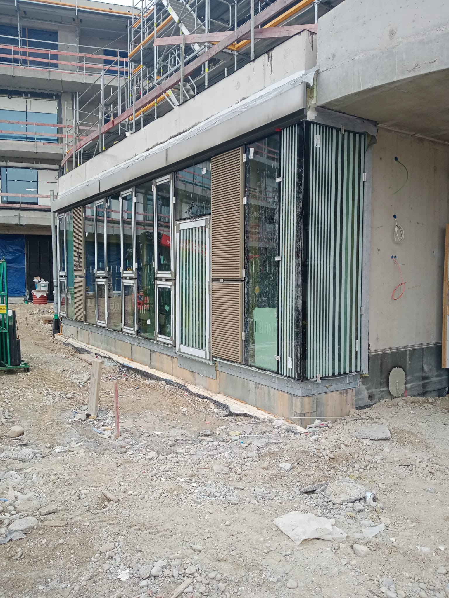 Wnętrze budynku w stanie surowym zamkniętym z zamontowanymi oknami fasadowymi i lamelami pionowymi, widok na plac budowy z gruzem i rusztowaniami w tle.